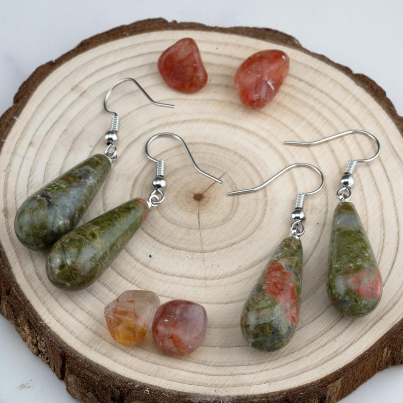 May include: Two pairs of drop earrings featuring polished, teardrop-shaped green and reddish-brown stone beads suspended from silver-toned metal hooks. Four loose, smooth, rounded reddish-brown stones are arranged around the earrings.