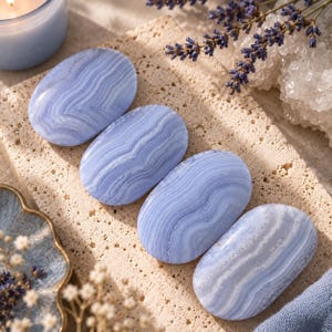 May include: Four smooth, oval-shaped blue lace agate stones featuring distinct white and light blue banded patterns. The polished gemstones are arranged in a diagonal line on a textured, porous beige stone surface alongside dried lavender.