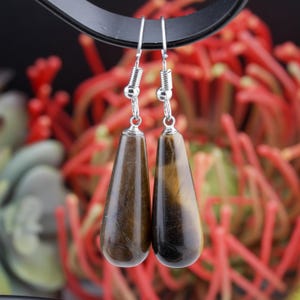May include: A pair of drop earrings featuring polished, teardrop-shaped tiger's eye gemstones in shades of brown and gold. The stones are suspended from silver-toned metal fishhook wires, showcasing a smooth, reflective surface and natural banding.