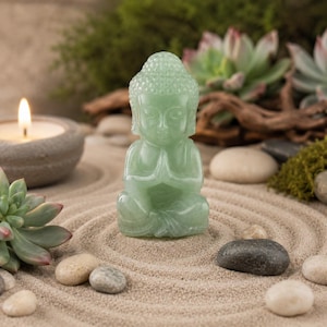May include: A serene scene featuring a small, light green Buddha statue in a meditative pose. The statue is set on a bed of sand, surrounded by small stones, succulents, and a lit candle in a gray holder. The overall aesthetic is calming and zen-like.