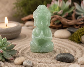 May include: A serene scene featuring a small, light green Buddha statue in a meditative pose. The statue is set on a bed of sand, surrounded by small stones, succulents, and a lit candle in a gray holder. The overall aesthetic is calming and zen-like.