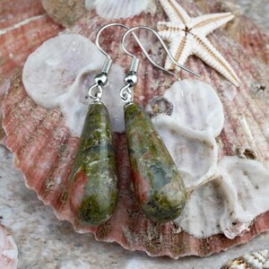 May include: A pair of teardrop-shaped earrings featuring polished unakite stone with mottled green and pink patterns. The stones are suspended from silver-toned metal fishhook wires, showcasing a smooth, glossy finish and natural stone variations.