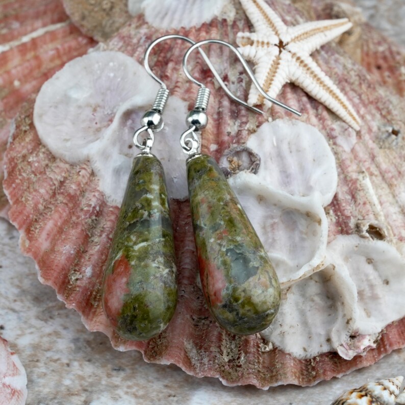 May include: A pair of teardrop-shaped earrings featuring polished unakite stone with mottled green and pink patterns. The stones are suspended from silver-toned metal fishhook wires, showcasing a smooth, glossy finish and natural stone variations.