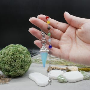 May include: A pendulum with a light blue crystal point and a silver-colored filigree cap. The pendulum is strung with a chain of colorful beads, including red, yellow, green, and blue. The background is dark, with a green plant and white stones.