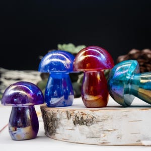 May include: Four iridescent glass mushroom figurines in shades of blue, red, purple, and green. The mushrooms are arranged on a white wooden surface.