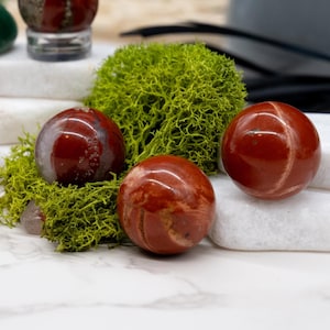 Puede incluir: Tres esferas pulidas y redondas de jaspe rojo, con vetas blancas y marrones, descansan sobre un lecho de musgo verde y piedra blanca. Las esferas tienen aproximadamente 5 cm de diámetro. Se ven esferas adicionales en el fondo.