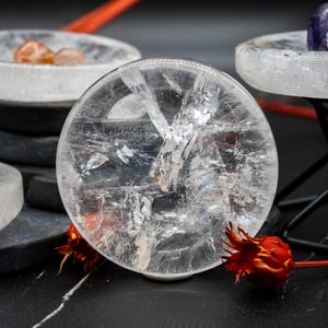 Pode incluir: Um disco circular de cristal de quartzo transparente polido, apresentando fraturas e inclusões internas naturais. A pedra transparente está posicionada na vertical, exibindo uma superfície lisa com facetas que refletem a luz por toda a estrutura.