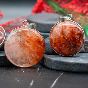 Op de afbeelding: Close-up van twee ronde hangers met een zilverkleurig metalen frame. De hangers hebben een rood-oranje, gemarmerde steen. De achtergrond bevat donkergrijze stenen en vage rode en groene decoraties, wat wijst op een vakantiethema.