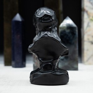 May include: A black bust sculpture with a textured surface, set against a backdrop of various crystal obelisks. The obelisks are in shades of blue, green, and white, creating a contrast with the dark sculpture.