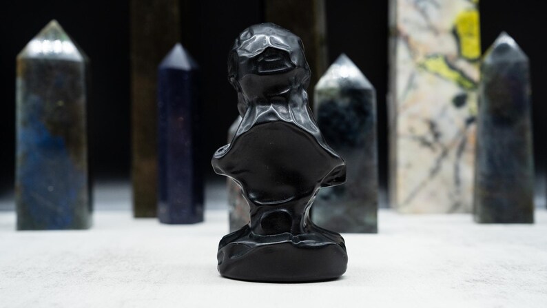 May include: A black bust sculpture with a textured surface, set against a backdrop of various crystal obelisks. The obelisks are in shades of blue, green, and white, creating a contrast with the dark sculpture.