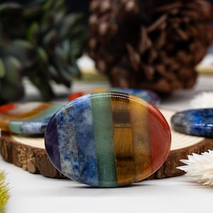 May include: A polished, oval-shaped stone with a rainbow of colors, including red, orange, yellow, green, blue, and purple. The stone is likely a chakra stone, used for meditation and healing. The stone is approximately 3 inches in diameter.