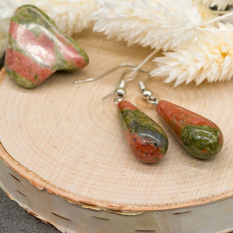 May include: A pair of teardrop-shaped unakite earrings featuring mottled green and pink stone patterns, suspended from silver-toned metal hooks, accompanied by a single polished unakite stone with a similar green and pink color palette.
