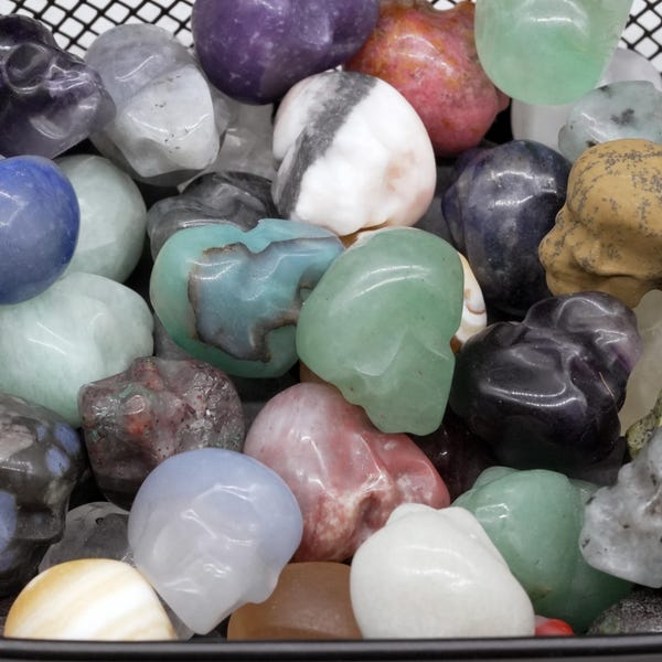 May include: A collection of small, polished gemstone carvings shaped like skulls. The assortment features various minerals in diverse colours, including shades of green, purple, blue, white, pink, brown, and black, with unique natural patterns and textures.