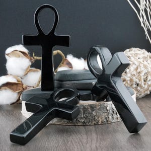 Può includere: Quattro simboli ankh neri lucidi sono esposti su una superficie di legno. Gli ankh, con la loro caratteristica parte superiore ad anello e forma a croce, sono disposti in gruppo, mostrando la loro finitura lucida e il design dettagliato. Lo sfondo include cotone ed elementi decorativi.