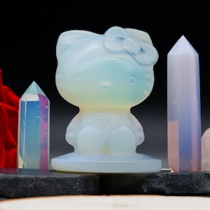 Puede incluir: Una colección de cristales y una figura de Hello Kitty opalescente blanca. Dos obeliscos de cristal y una rosa roja se presentan sobre una superficie oscura contra un fondo negro.