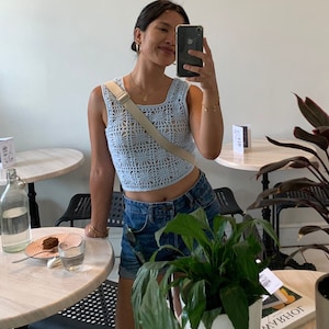 Op de afbeelding: Een lichtblauw gehaakt topje met een vierkante halslijn. De top wordt gecombineerd met een spijkerbroek. Een beige crossbodytas wordt over het lichaam gedragen. De foto is gemaakt in een café met ronde tafels en planten.