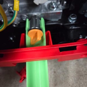 May include: A light green plastic oil drain funnel positioned beneath a metallic engine component. The funnel features a bright orange twist-cap valve and is set against a red plastic frame, with visible electrical wiring and metallic bolts nearby.