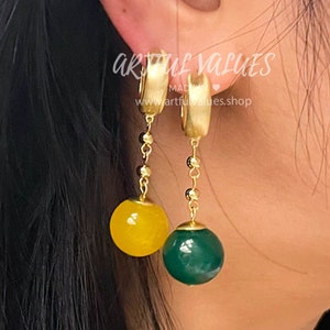 Könnte beinhalten: Goldene Creolen mit einem baumelnden grünen und gelben Stein. Die Ohrringe werden von der Firma "artful values" hergestellt.