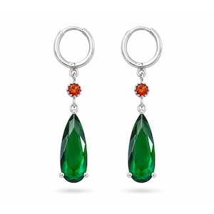 Puede incluir: Un par de pendientes de aro plateados con una pequeña piedra preciosa redonda de color naranja suspendida sobre una piedra más grande en forma de pera de color verde oscuro. Los pendientes tienen un acabado metálico pulido.