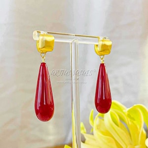 Könnte beinhalten: Ein Paar goldfarbene Ohrringe mit roten tropfenförmigen Steinen. Die Ohrringe befinden sich auf einem Ausstellungsständer.