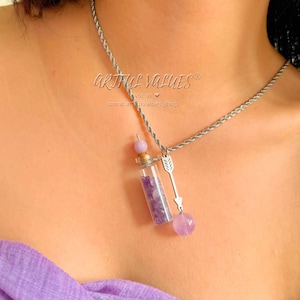 May include: A silver chain necklace with a small glass vial filled with purple amethyst crystals. The vial is attached to a silver arrow charm and a larger purple amethyst bead.