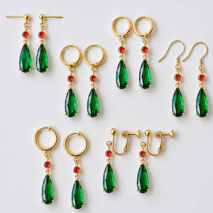 Puede incluir: Una colección de cinco pares de pendientes dorados que presentan una piedra preciosa verde en forma de lágrima suspendida bajo una pequeña piedra roja redonda. El conjunto incluye varios estilos de pendientes como broches, aros, ganchos y clips.