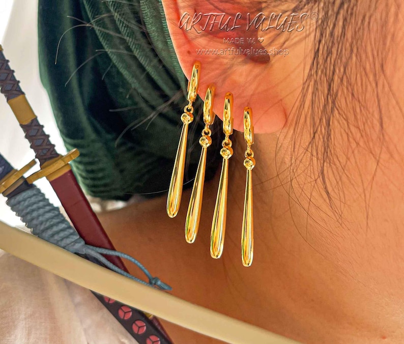 Peut inclure: Boucles d'oreilles pendantes dor&eacute;es en forme de larme. Les boucles d'oreilles sont port&eacute;es &agrave; l'oreille d'une personne. Les boucles d'oreilles sont fabriqu&eacute;es par Artful Values.