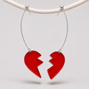 Puede incluir: Un par de pendientes colgantes que presentan dos mitades de corazón de acrílico rojo translúcido. Cada pieza de corazón está suspendida de un fino alambre metálico curvado de tono plateado que se conecta a un pequeño poste de bola plateada.