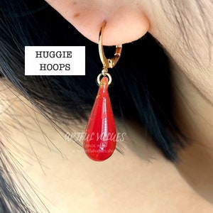 Pu&ograve; includere: Orecchino a cerchio dorato con un ciondolo rosso a forma di goccia. Il ciondolo ha un design sfaccettato e sembra essere fatto di un materiale traslucido. Il testo "HUGGIE HOOPS" &egrave; in un riquadro bianco.
