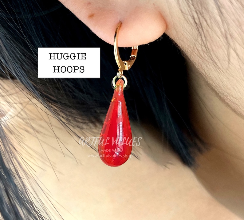 Pu&ograve; includere: Orecchino a cerchio dorato con un ciondolo rosso a forma di goccia. Il ciondolo ha un design sfaccettato e sembra essere fatto di un materiale traslucido. Il testo "HUGGIE HOOPS" &egrave; in un riquadro bianco.