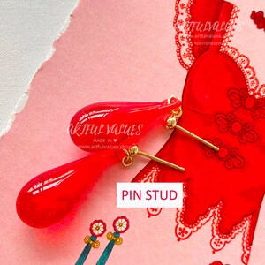 Pu&ograve; includere: Un paio di orecchini a perno a forma di goccia rossi con perni color oro. Gli orecchini sono esposti su una superficie rosa con un'illustrazione di un vestito rosso sullo sfondo. Il testo "PIN STUD" &egrave; visibile.