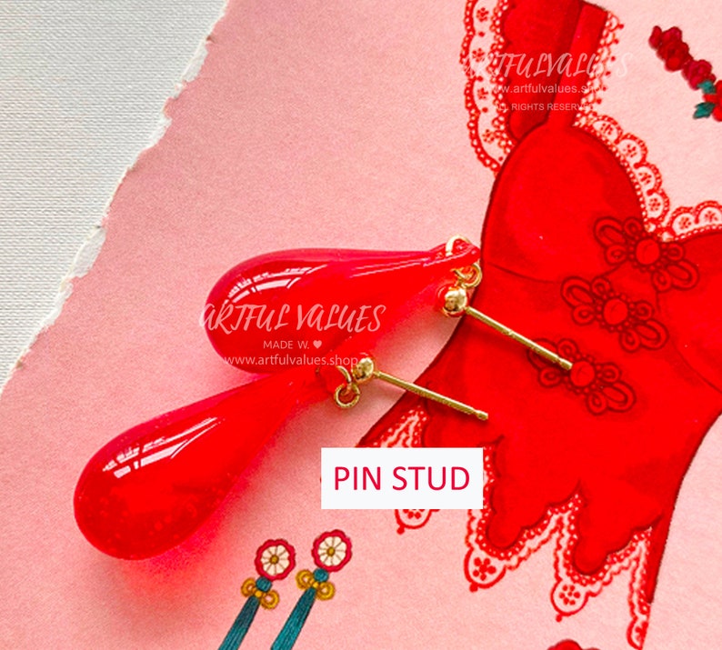 Pu&ograve; includere: Un paio di orecchini a perno a forma di goccia rossi con perni color oro. Gli orecchini sono esposti su una superficie rosa con un'illustrazione di un vestito rosso sullo sfondo. Il testo "PIN STUD" &egrave; visibile.