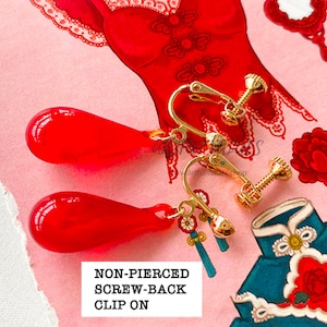Pu&ograve; includere: Orecchini rossi a goccia con chiusura a clip e elementi dorati. Gli orecchini hanno una clip a vite e piccoli nappe decorative. Lo sfondo presenta illustrazioni di abiti tradizionali cinesi e motivi floreali.