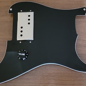 May include: Black guitar pickguard with a silver pickup cover and a volume knob. The pickguard has a white outline and is made of plastic.
