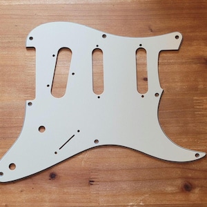 May include: Off-white guitar pickguard with three elongated pickup cutouts and various screw holes. The pickguard has a curved shape with a pointed lower edge and a small extension on the upper edge. The pickguard is made of a solid, opaque material.
