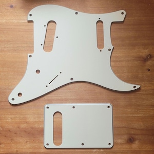 Peut inclure: Un ensemble blanc de plaque de protection et de cache trémolo pour guitare. La plaque de protection a une forme profilée avec des trous de vis et des découpes pour les micros et les commandes. Le cache trémolo est rectangulaire avec une seule découpe.