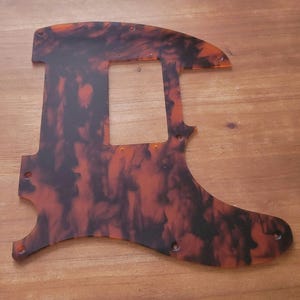 Puede incluir: Un golpeador de guitarra de caparazón de tortuga negro y naranja con un recorte para una pastilla y varios agujeros para tornillos.