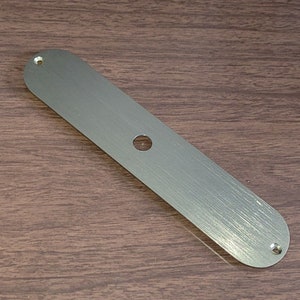 Könnte beinhalten: Eine gebürstete silberne Metallplatte mit abgerundeten Kanten und zwei Schraubenlöchern. Die Platte hat ein einzelnes rundes Loch in der Mitte.