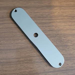 May include: A polished chrome guitar control plate. The plate is a long, rectangular shape with rounded ends. It has three holes: one central and one at each end. The plate reflects the surrounding environment.