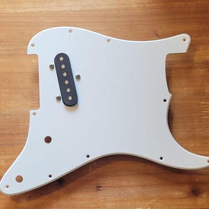 Op de afbeelding: Een witte gitaar slagplaat met een zwarte pickup en bevestigingsschroeven. De slagplaat heeft een gebogen vorm met verschillende gaten voor bevestiging aan een gitaarbody. Dit gitaar onderdeel is voor elektrische gitaren.