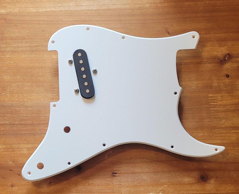Op de afbeelding: Een witte gitaar slagplaat met een zwarte pickup en bevestigingsschroeven. De slagplaat heeft een gebogen vorm met verschillende gaten voor bevestiging aan een gitaarbody. Dit gitaar onderdeel is voor elektrische gitaren.