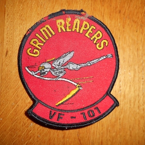May include: A circular red patch with the words "GRIM REAPERS" in yellow lettering. The patch features a skeleton figure with a scythe. The text "VF-101" is at the bottom.