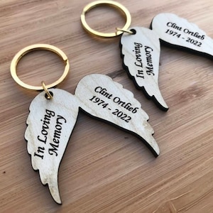 Può includere: Due portachiavi in legno a forma di ali d'angelo con anelli portachiavi dorati. Le ali sono incise con "In Loving Memory" e "Clint Ortlieb 1974 - 2022".