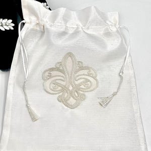 Può includere: Borsa a sacco in raso bianco con un design a forma di giglio d'argento ricamato e lacci con nappe.