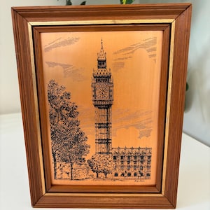 May include: A rectangular wall art piece featuring a copper-toned metal plate with a black etched illustration of the Big Ben clock tower and surrounding trees. The artwork is encased in a dark brown wooden frame with a thin gold-colored inner trim.