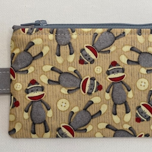 Può includere: Una piccola pochette con cerniera, con una cerniera grigia e una tracolla grigia con un portachiavi. La pochette è realizzata in tessuto beige con un motivo di scimmie di calzini grigi che indossano cappelli rossi.