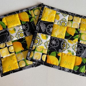 May include: Two square quilted pot holders with a patchwork design. The design features yellow lemons, lemon slices, and floral patterns on a black and white background. The edges are black with a white floral pattern.
