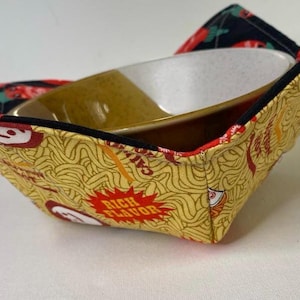May include: A yellow fabric bowl cozy with a ramen noodle pattern and the words "Rich Flavor" in a red burst. A ceramic bowl sits inside the cozy. The rim of the cozy is black, and the interior is a black and red pattern.