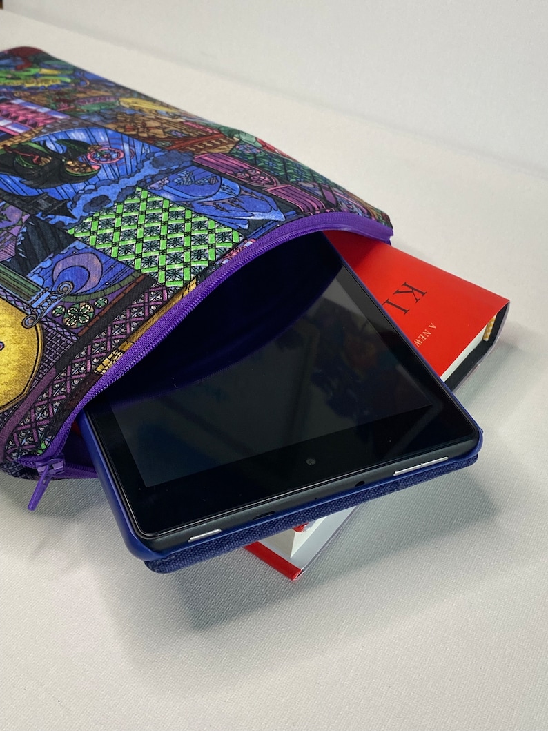 K&ouml;nnte beinhalten: Eine violette Rei&szlig;verschlusstasche mit einem bunten Buntglasmuster. Die Tasche ist offen und ein schwarzes Tablet-Computer befindet sich darin. Das Tablet liegt auf einem roten Buch mit dem sichtbaren Text "A YEAR" und "XII".