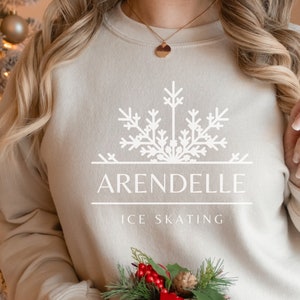 K&ouml;nnte beinhalten: Ein beiger Pullover mit einem wei&szlig;en Schneeflocken-Design und dem Text "Arendelle Ice Skating". Die Person h&auml;lt eine rote und schwarze karierte Geschenkbox mit einem gr&uuml;nen Zweig und roten Beeren.